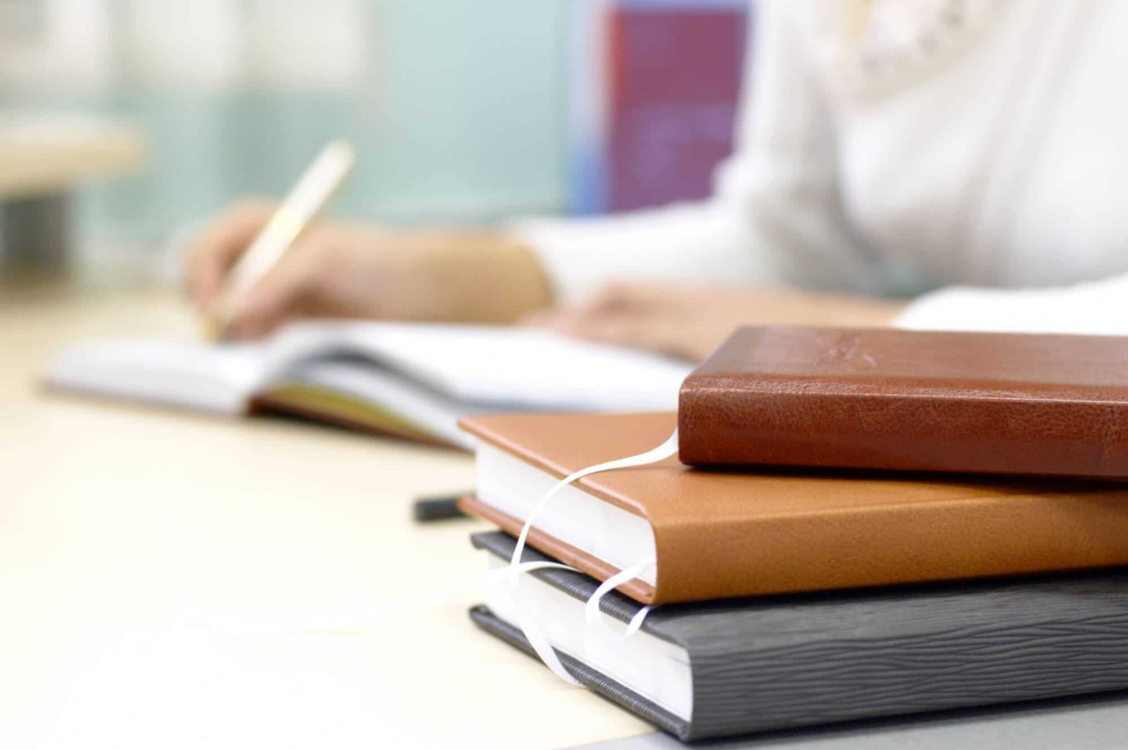 Imagem de 3 livros empilhados em uma mesa, com o braço de uma pessoa ao fundo desfocada, se apoiando na mesa e escrevendo em um caderno.